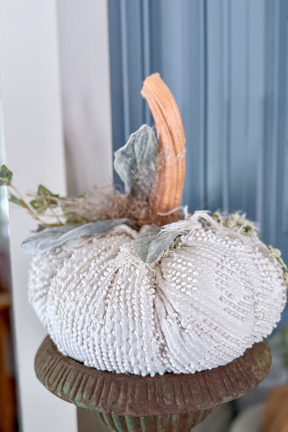 A decorative white fabric pumpkin with a textured surface, faux leaves, and a brown stem sits on a rustic pedestal in front of a blue paneled wall—perfect inspiration if you’re learning how to make fabric pumpkins.
