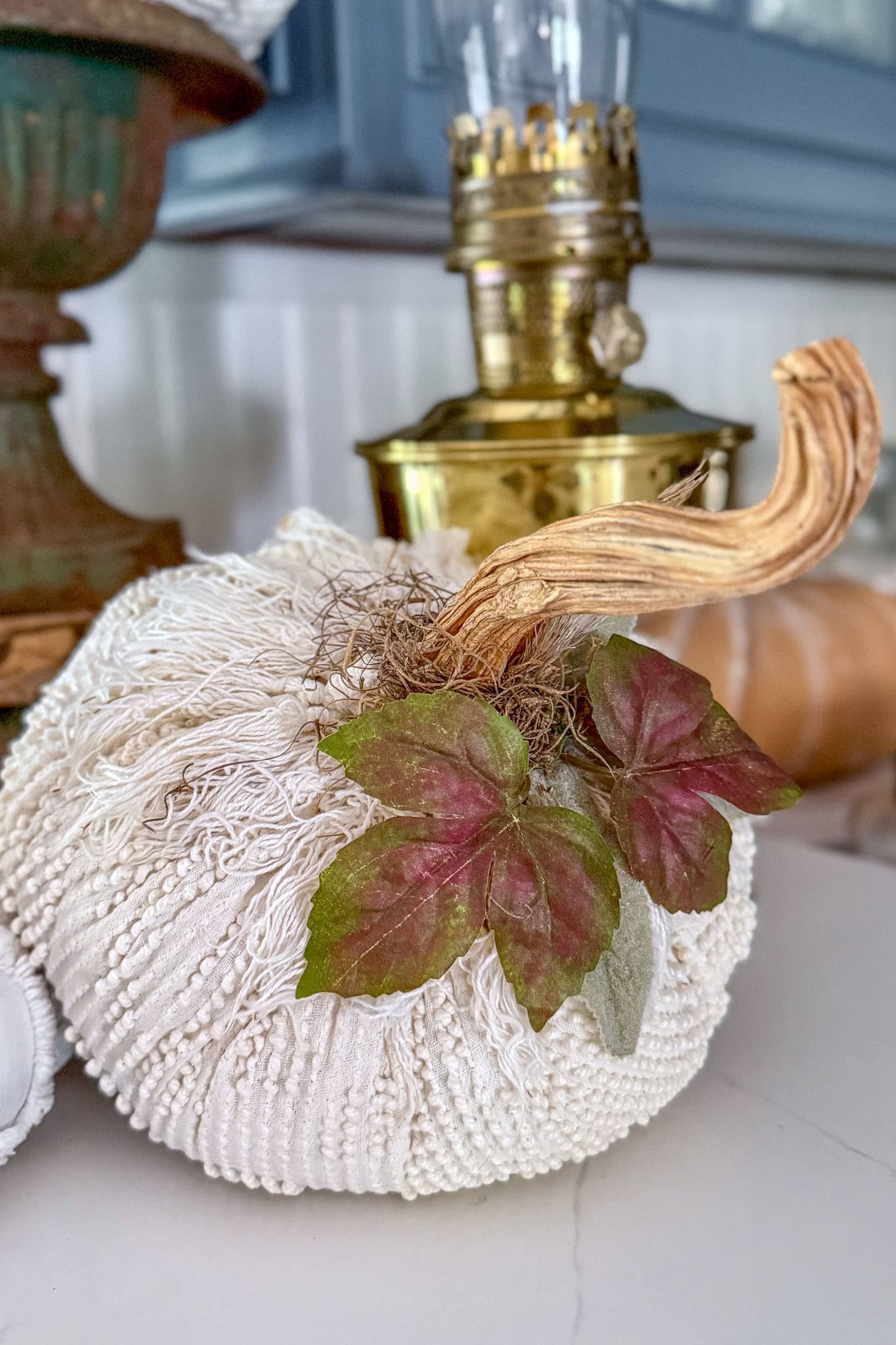 A decorative pumpkin, perfect for those learning how to make fabric pumpkins, crafted from textured white fabric with a twisted stem, two green and reddish leaves, and a touch of moss, sits on a table beside a brass lamp.