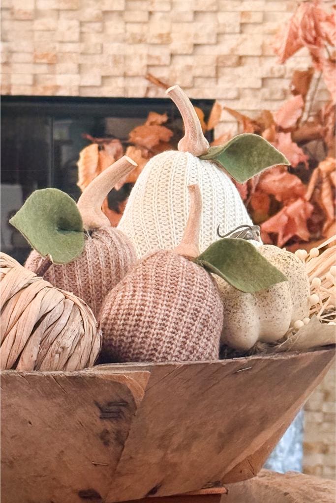A wooden bowl filled with decorative fabric pumpkins in beige, cream, and brown tones, with green felt leaves. In the background, autumn leaves and a brick wall are visible.
