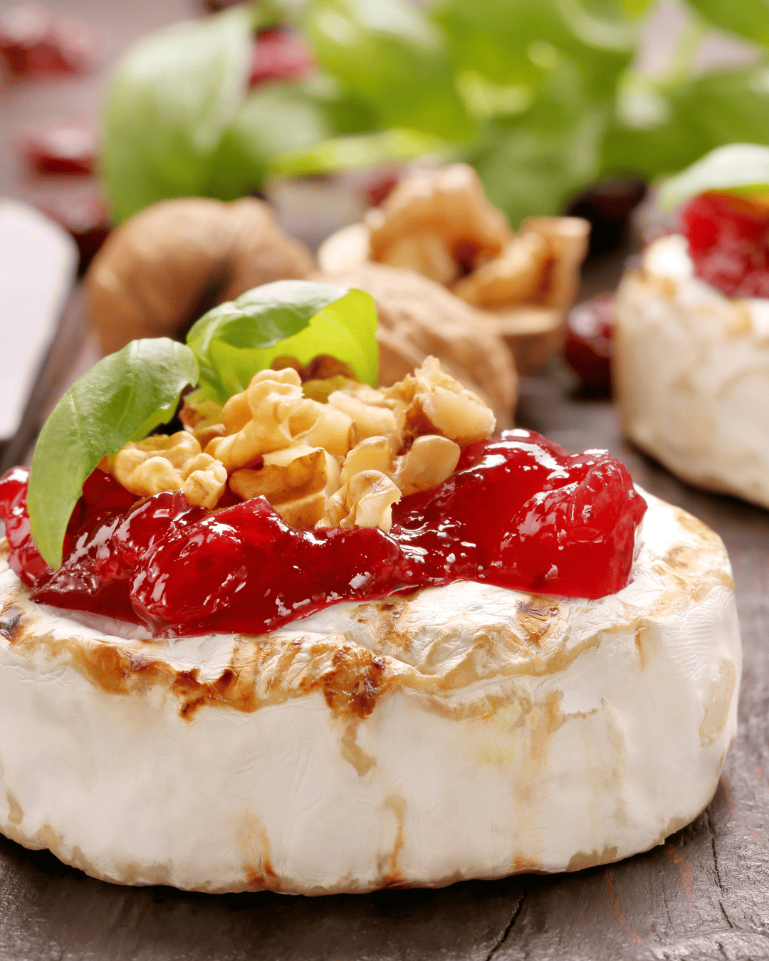 A round of brie cheese topped with red fruit jam, chopped walnuts, and fresh basil leaves, with walnuts and greens blurred in the background—perfect for pairing with an Easy Cherry Tomato Jam Recipe for Beginners.