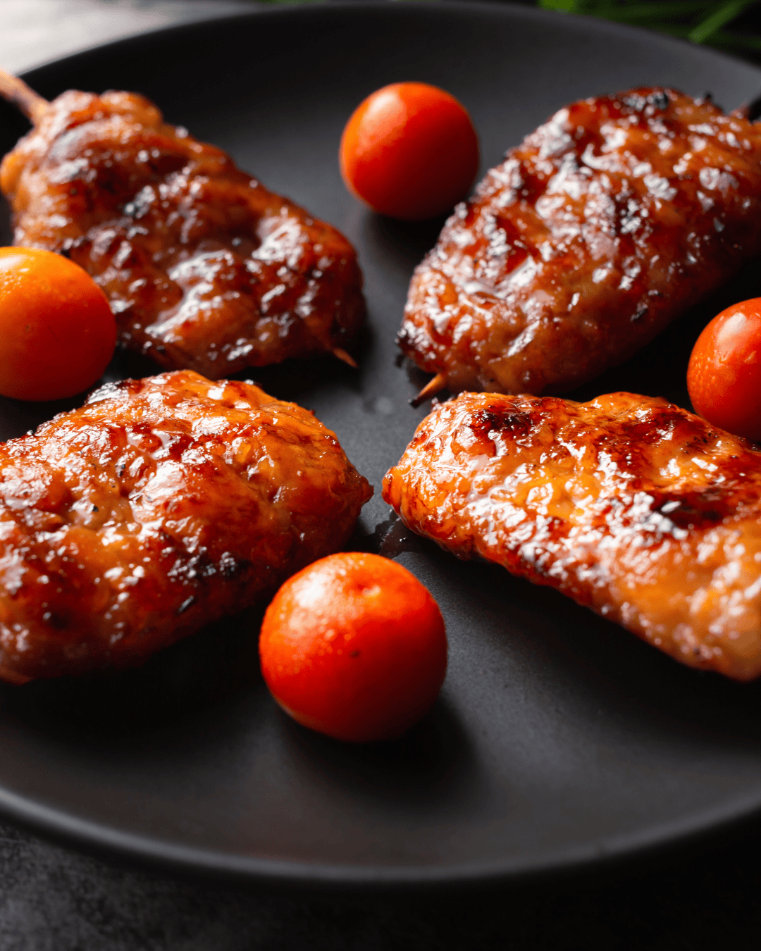 Grilled chicken pieces with a glossy glaze are arranged on a dark plate, surrounded by four bright red cherry tomatoes, evoking the fresh flavors celebrated in any Easy Cherry Tomato Jam Recipe for Beginners.