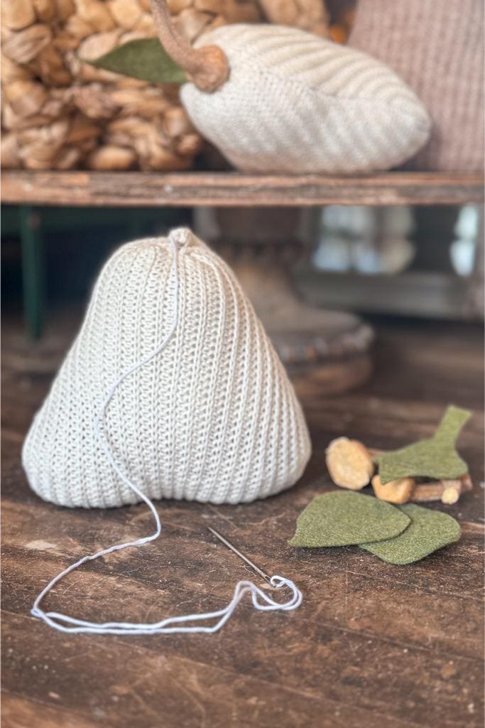 DIY Sweater Pears-A knitted pear-shaped object with a needle and white thread attached sits on a wooden surface. Felt leaf pieces and stuffing are nearby, and another finished knit pear is in the background.