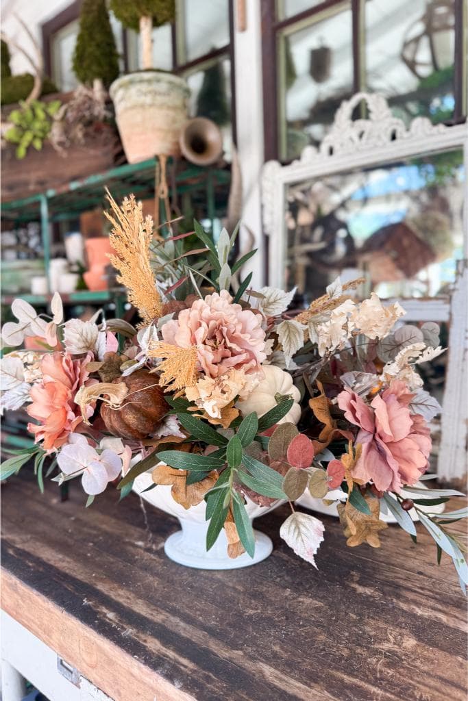 budget-friendly DIY fall centerpiece with pumpkins and greenery-A white vase filled with a large arrangement of pastel and dried flowers sits on a rustic wooden table; a mirror and potted plants are in the background, creating a cozy, vintage atmosphere.