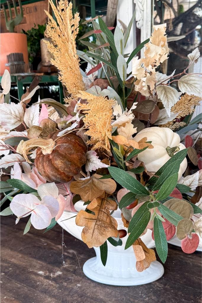 A white ceramic vase filled with an autumn floral arrangement, featuring dried leaves, grasses, green foliage, and two pumpkins—one white and one brown—resting on a wooden table.
