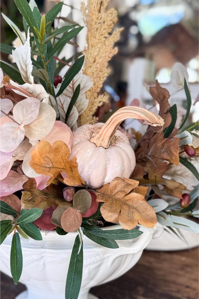 A white ceramic bowl filled with autumn-themed decor, featuring a small, pale pumpkin, dried leaves, green foliage, and pinkish flowers.