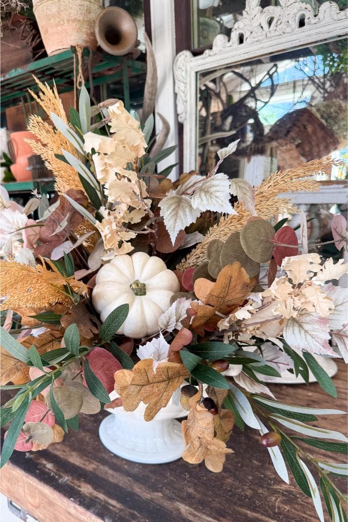 A white ceramic vase holds a fall arrangement of dried leaves, grasses, and foliage, with a small white pumpkin in the center, displayed on a rustic wooden table with a mirror and plants in the background.