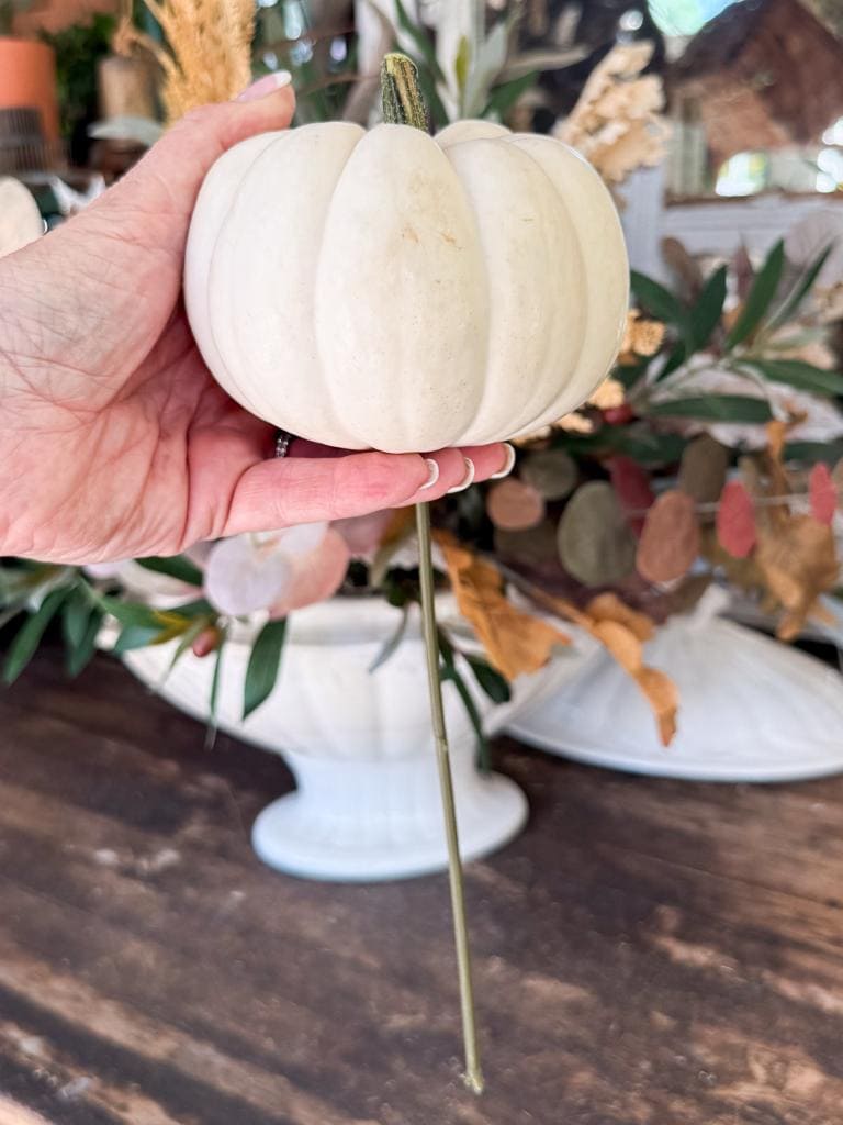A hand holds a small white pumpkin attached to a long green stem, with a floral arrangement and white ceramic bowl on a wooden table in the background.