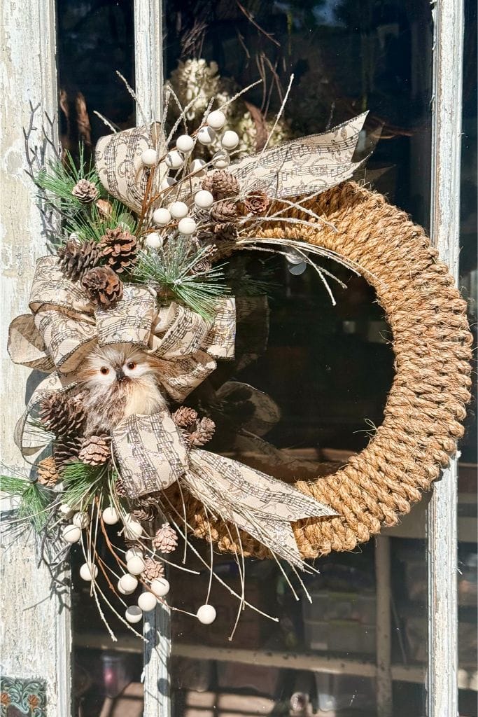 A rustic wreath made of braided rope hangs on a door, adorned with pinecones, white berries, greenery, sheet music ribbon, and a small artificial owl for decoration. Sunlight highlights its natural texture.