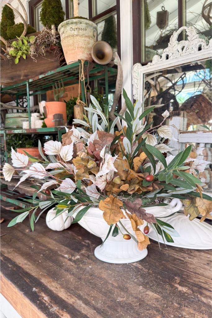 A white vase holds an arrangement of autumn leaves and greenery on a wooden table, surrounded by white ceramic pumpkins. Garden pots and tools are visible in the background on green metal shelves.
