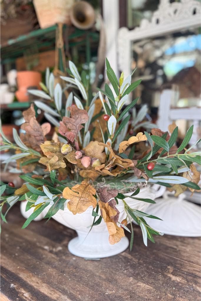 A white ceramic vase holds an arrangement of green and brown leaves and dried foliage, set on a rustic wooden table with a blurred background featuring shelves, pots, and a decorative mirror.