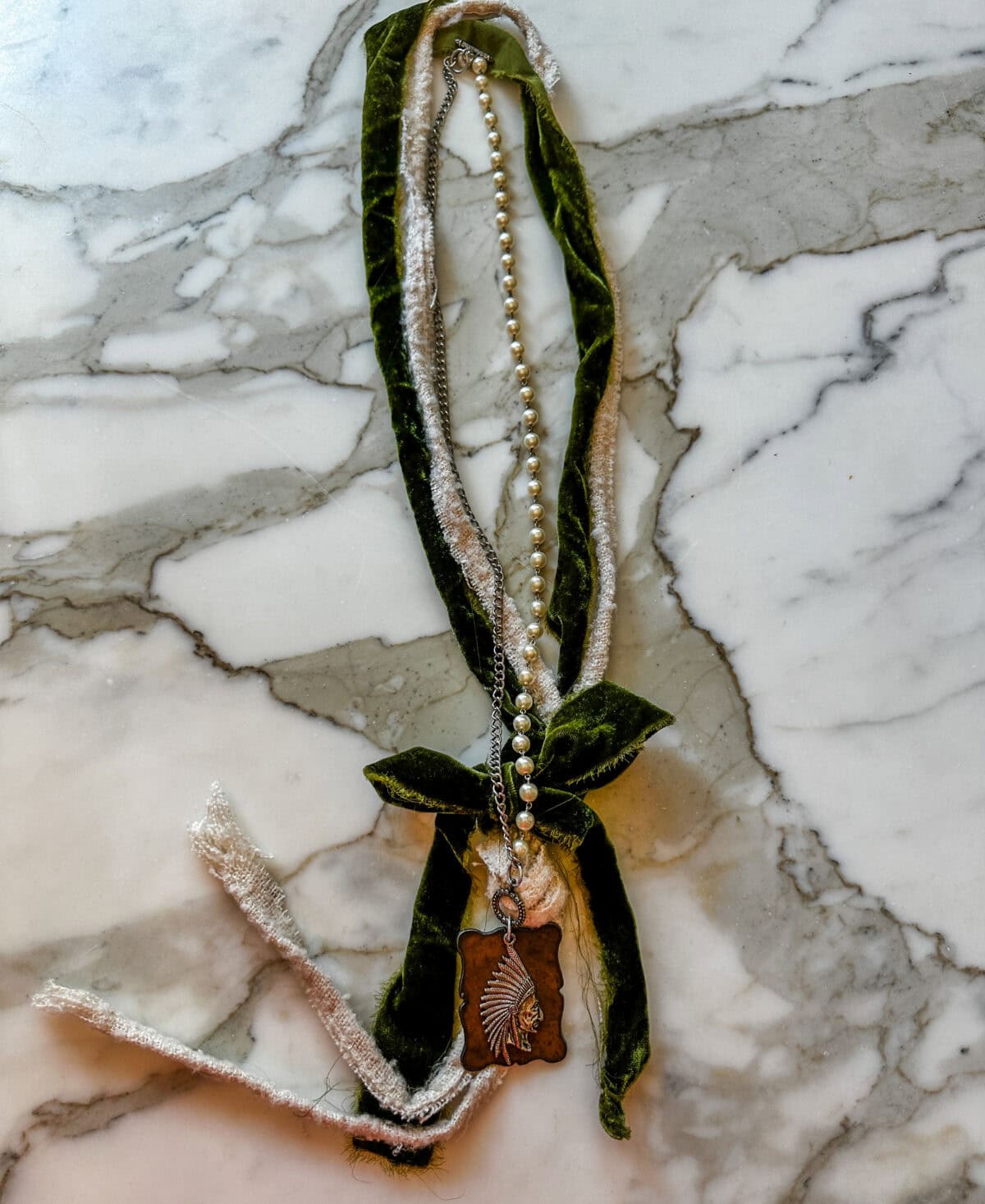 A necklace with a strand of pearls and green velvet ribbon, tied in a bow, featuring a rectangular pendant engraved with a leaf design, lies on a white marble surface.
