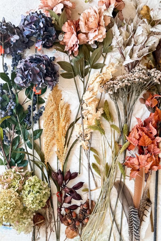 A flat lay of assorted artificial flowers and foliage, including hydrangeas, peonies, pampas grass, autumn leaves, berries, and feathers, arranged neatly on a light fabric background.