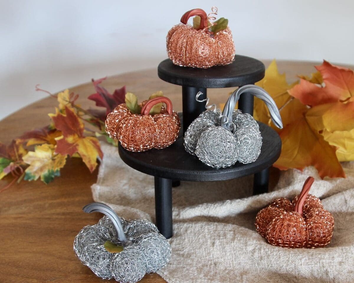 Five decorative pumpkins made of copper and silver scrubbers sit on and around a small black stand, with autumn leaves scattered in the background on a wooden surface—a charming display of pumpkin crafts for autumn décor.