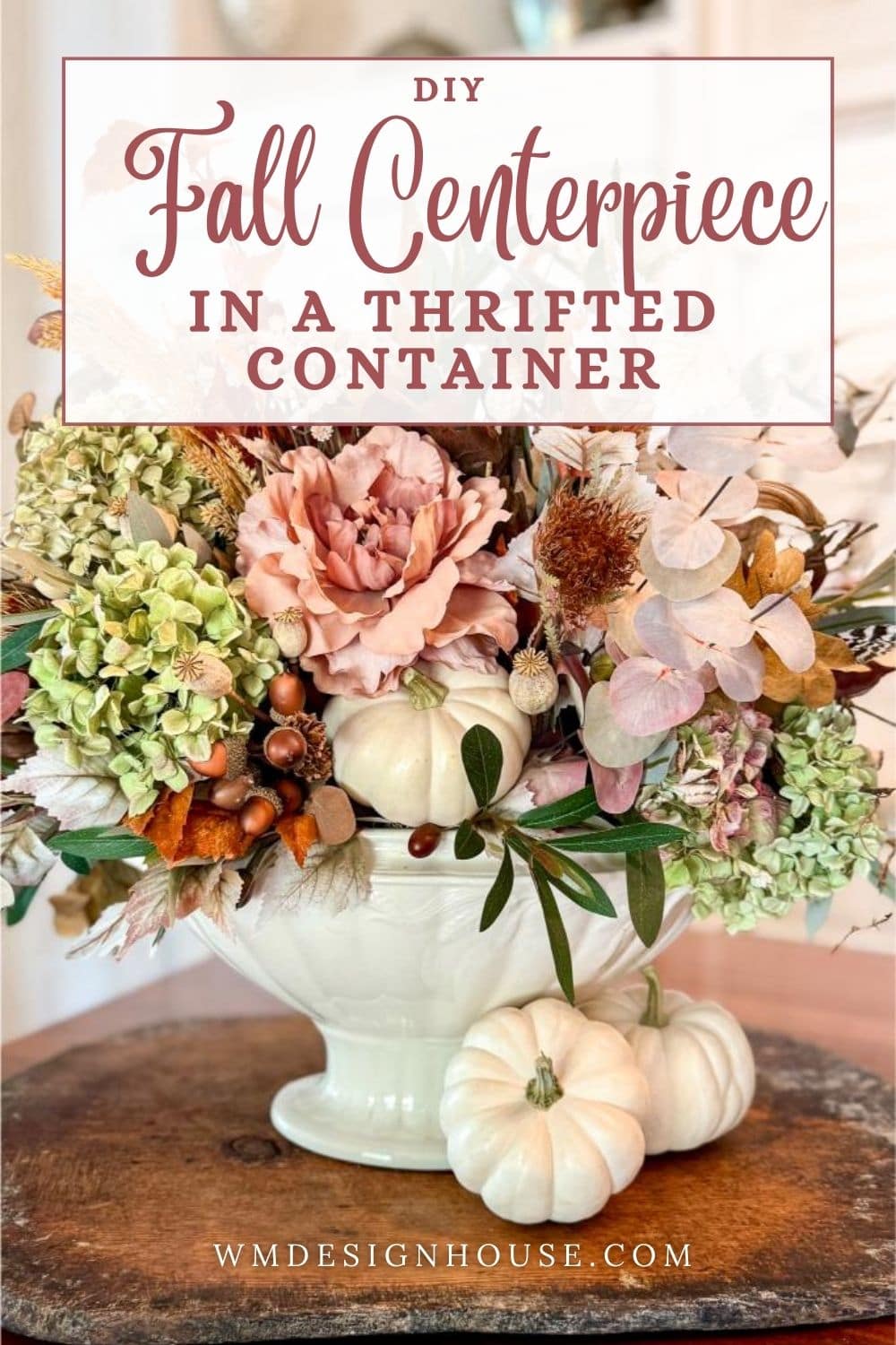 A DIY fall centerpiece with thrifted container features pink, green, and cream flowers with leaves and dried accents in a white vase, paired with two small white pumpkins on a wooden table. Text reads “DIY Fall Centerpiece in a Thrifted Container.”.