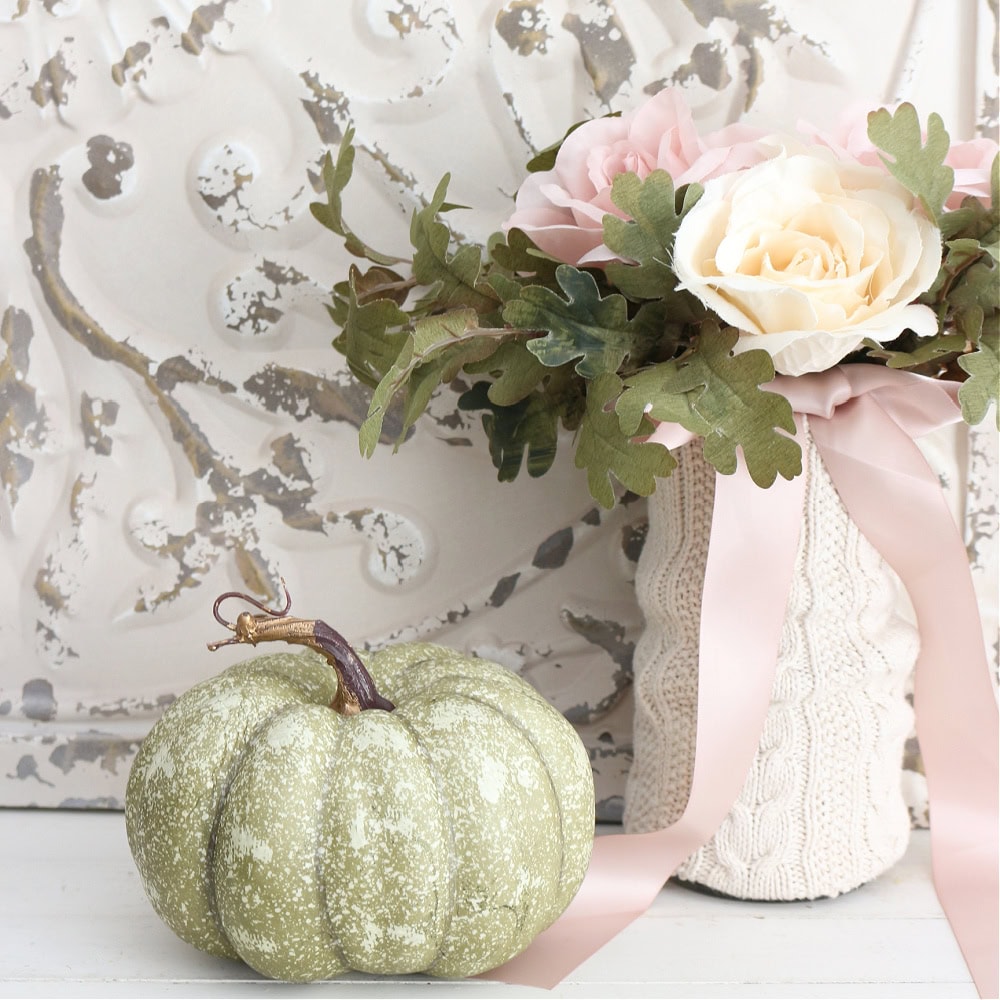 A pale green pumpkin sits beside a vase wrapped in a knitted cover, filled with pink and white roses and green leaves, set against a decorative white background.