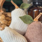 Three DIY Sweater Pears with knitted textures and felt leaves are displayed together. The pears, in cream, light brown, and beige, are set against a blurred background with a woven basket and glass bottle.