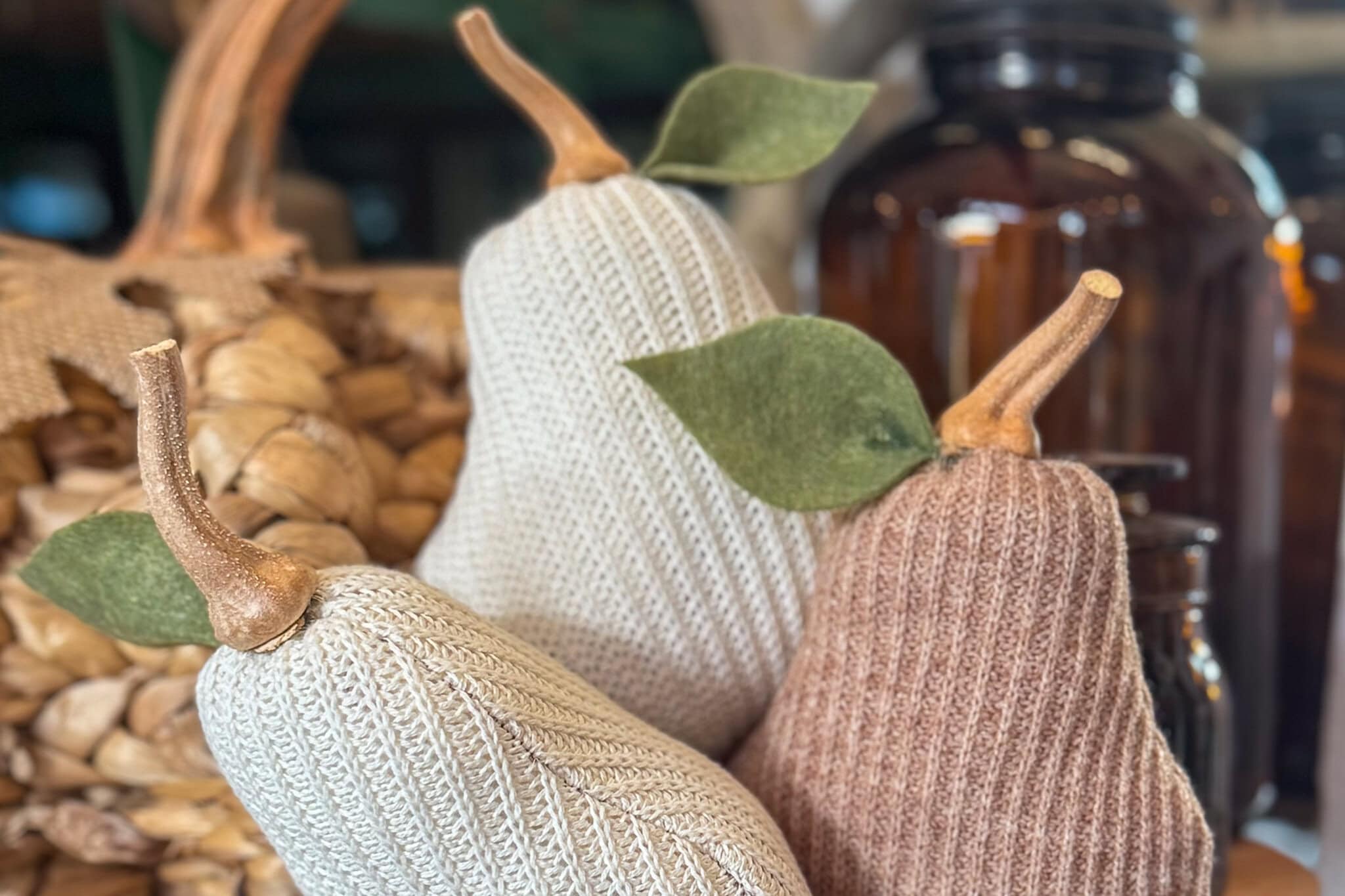 Three DIY Sweater Pears with knitted textures and felt leaves are displayed together. The pears, in cream, light brown, and beige, are set against a blurred background with a woven basket and glass bottle.