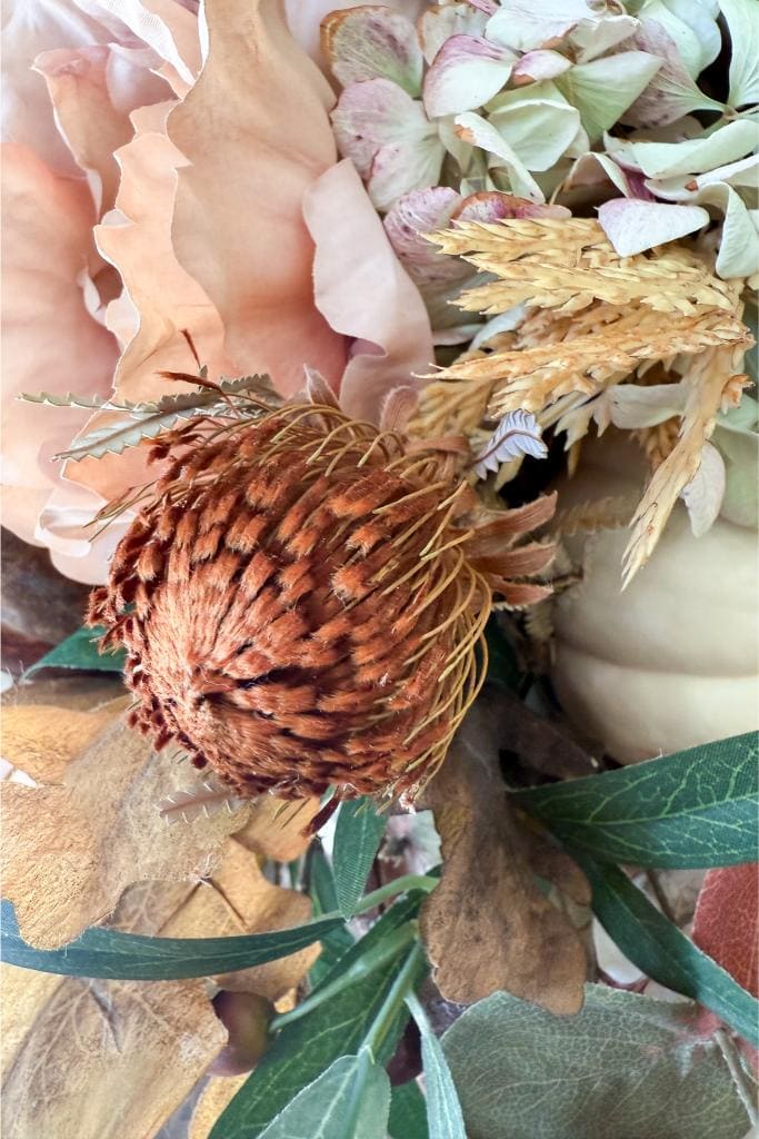 A close-up of a dried floral arrangement featuring a large brown protea flower, beige wheat stems, muted pink petals, and green leaves. The overall colors are earthy and soft, creating a rustic look.