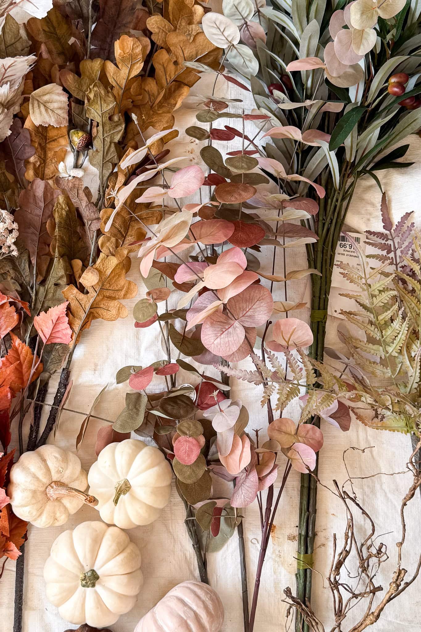 A variety of autumn-themed artificial leaves, ferns, and stems are arranged with small white decorative pumpkins on a light fabric background—perfect for a DIY fall centerpiece with thrifted container in muted pinks, greens, browns, and creams.