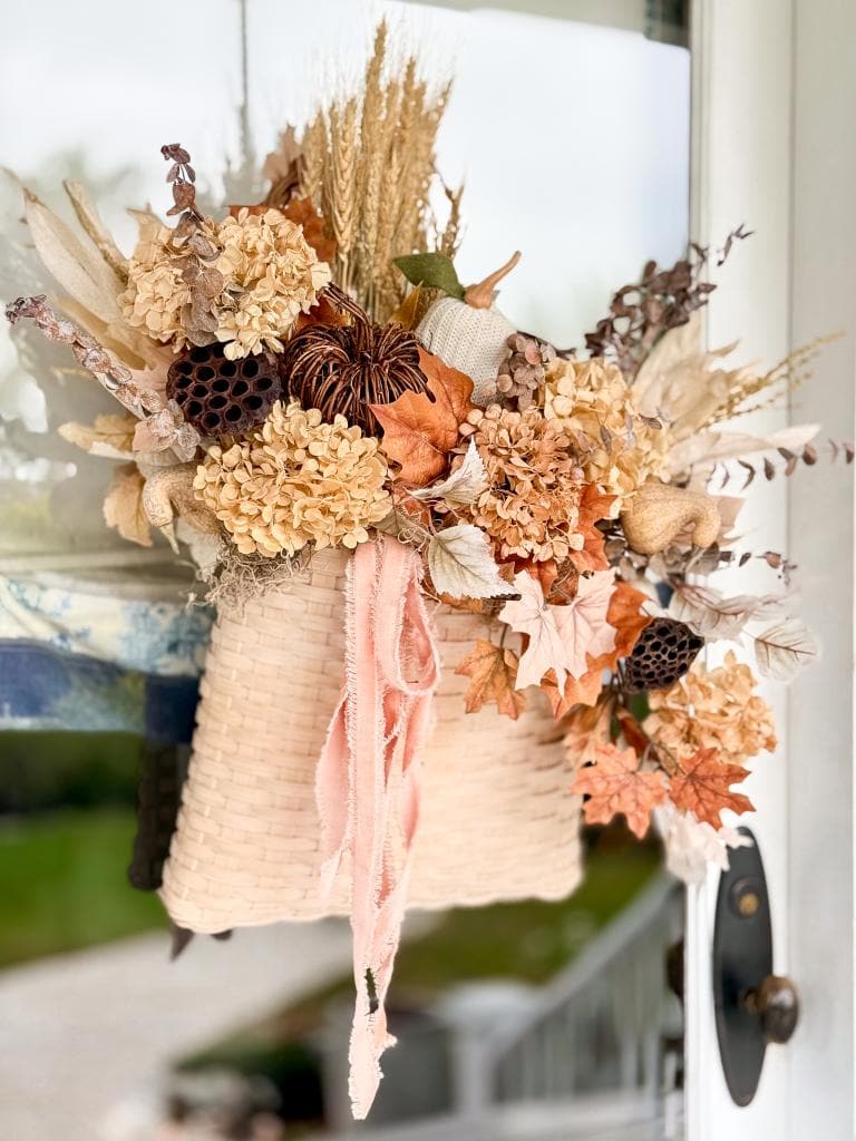 A woven basket filled with dried flowers, leaves, and autumn foliage hangs on a door, decorated with soft pink ribbons for a fall-themed front door display.