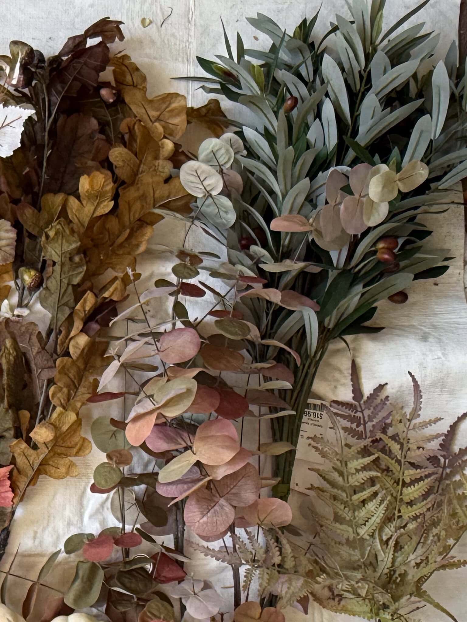 A variety of dried and preserved leaves, including eucalyptus, oak, and fern, are arranged on a cream-colored fabric background—perfect for creating a DIY fall centerpiece with a thrifted container. The leaves display muted green, brown, and mauve tones.