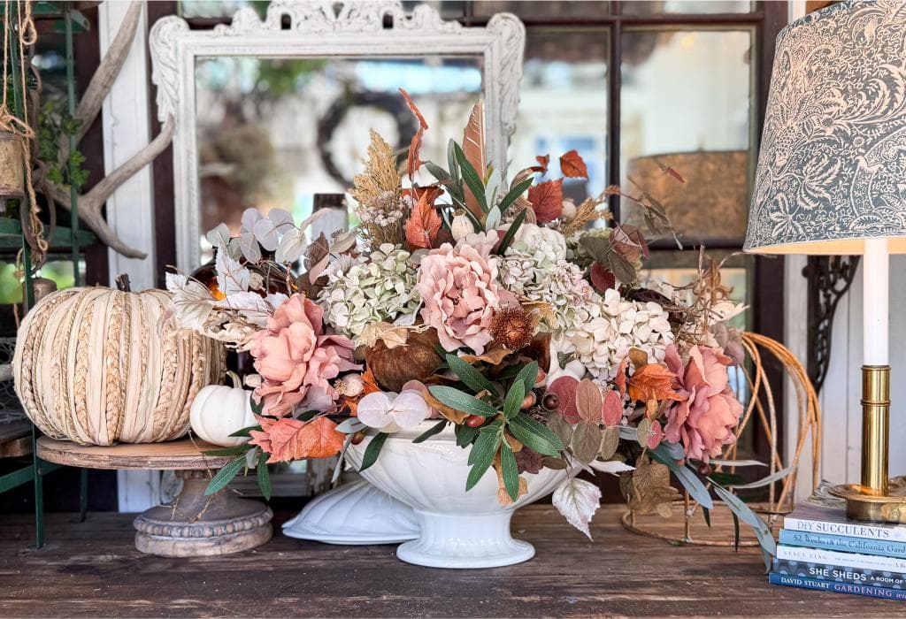 A large floral arrangement with pink, white, and brown flowers and leaves sits in a white bowl on a wooden table, next to a decorative pumpkin, stacked books, and a lamp with a patterned shade. A mirror is in the background.