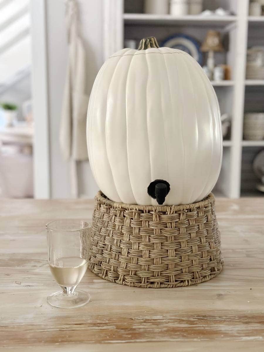 A white pumpkin-shaped drink dispenser, perfect for Pumpkin Crafts enthusiasts, sits on a woven basket base. A glass of light-colored beverage rests beside it on a wooden table, with shelves of dishes in the background.