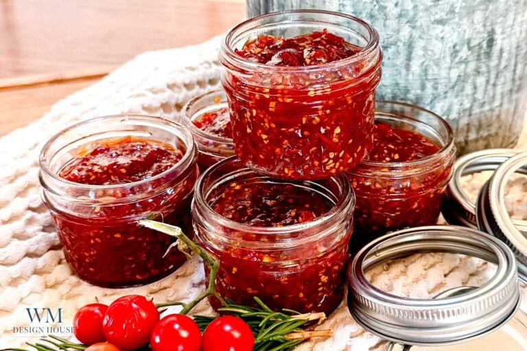 Five small glass jars filled with red tomato jam are stacked on a textured cloth. Open jar lids and fresh cherry tomatoes with rosemary are arranged beside the jars, capturing the inviting look of an Easy Cherry Tomato Jam Recipe for Beginners.