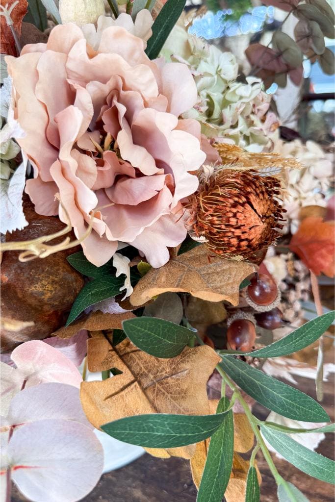 A close-up of an autumn-themed bouquet with a large pale pink flower, dried brown leaves, green foliage, and a spiky brown seed pod among other natural elements.