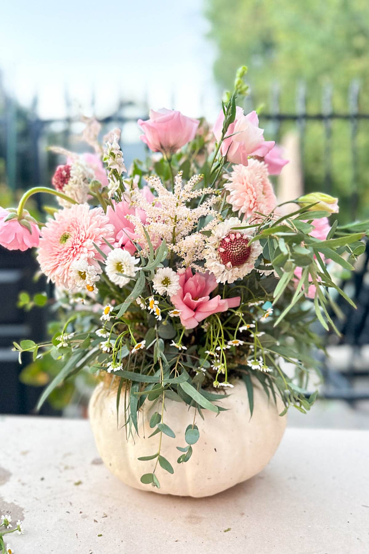 A white pumpkin is used as a vase for this charming Pumpkin Crafts display, holding an arrangement of pink and white flowers, greenery, and small daisies, set outdoors with a blurred fence and trees in the background.