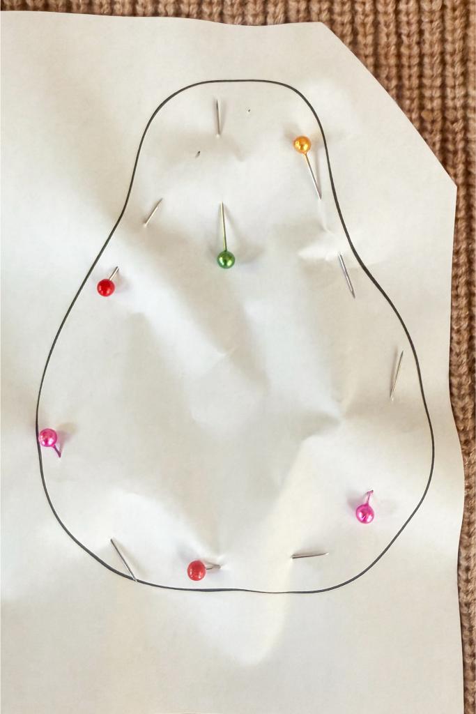 DIY Sweater Pears-A roughly pear-shaped piece of white paper with a black outline lies on a textured brown surface. Colorful pins, including red, pink, green, orange, and white, are pinned along the outline.