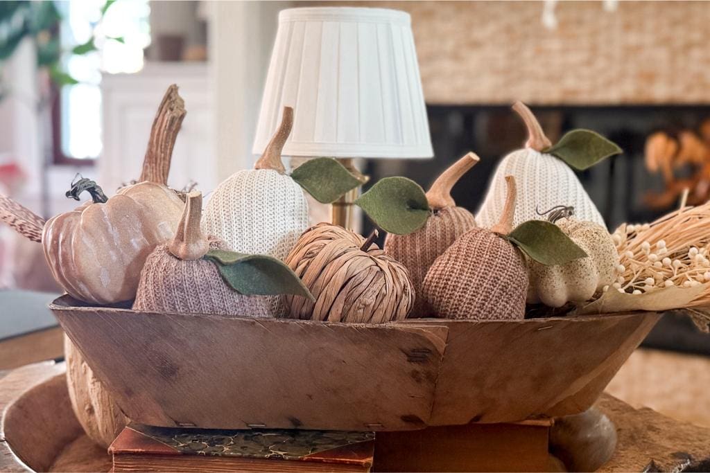 DIY Sweater Pears-A wooden bowl filled with decorative pumpkins made of various materials like yarn, fabric, and wicker sits on a table; a small lamp and fireplace are blurred in the background.