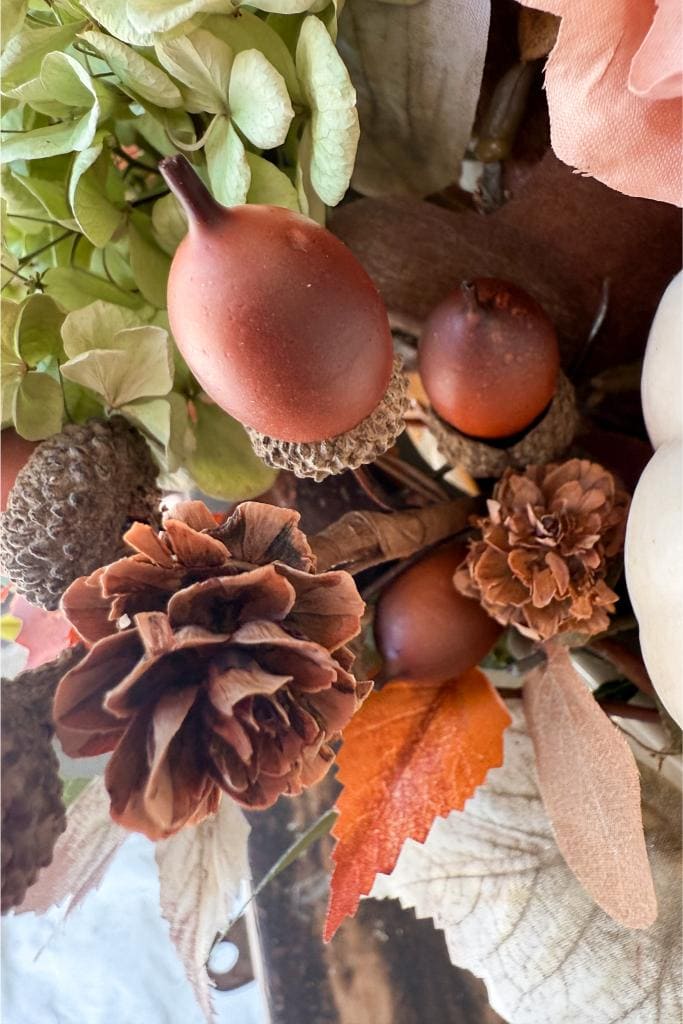 A close-up of an autumn arrangement featuring pinecones, acorns, orange and cream leaves, dried flowers, and green foliage. The colors are earthy, including browns, oranges, creams, and greens.