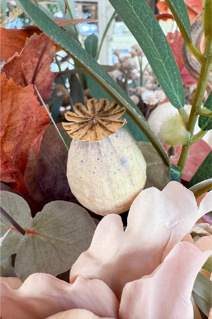 A dried poppy seed pod surrounded by artificial leaves and pastel pink fabric flowers in a decorative arrangement.