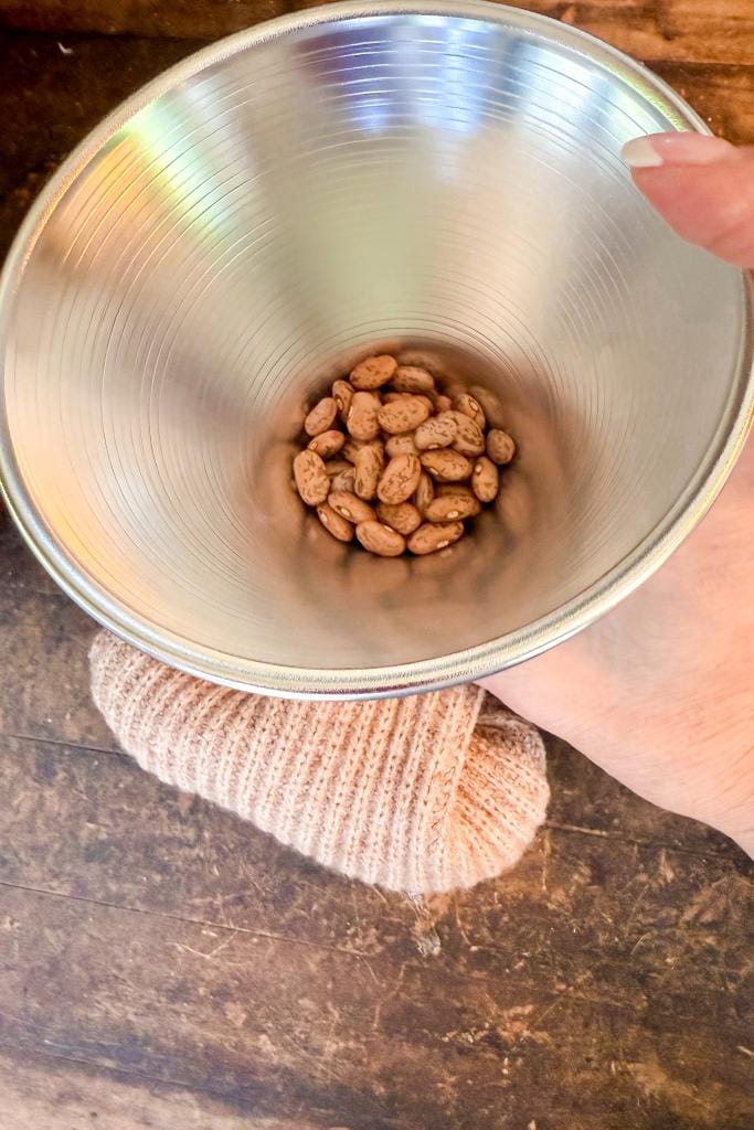 DIY Sweater Pears-A hand holding a shiny metal bowl containing a small pile of pinto beans, with a beige knit cloth on a wooden surface below.