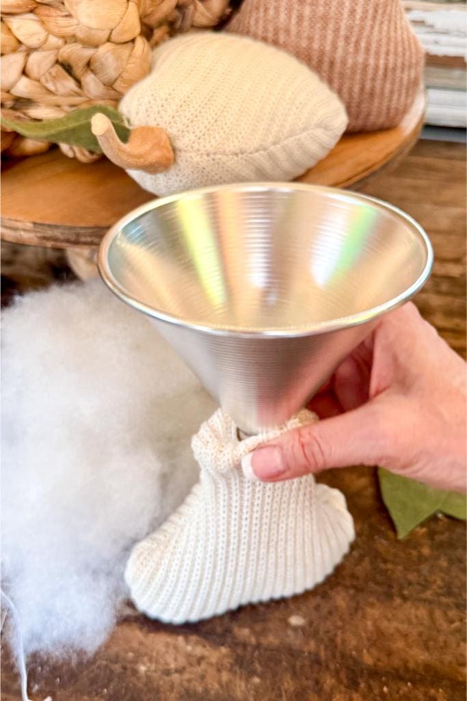 DIY Sweater Pears-A hand holds a metal funnel placed into a small, white, knitted sock, with synthetic stuffing and decorative fabric pumpkins on a wooden surface in the background.
