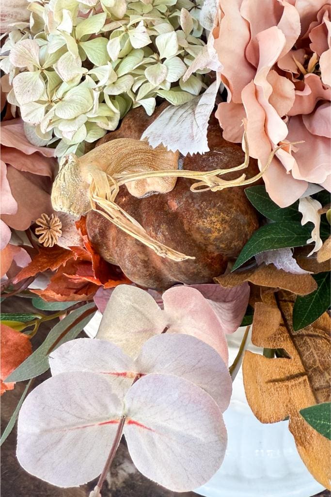 A close-up of a floral arrangement featuring pale green hydrangeas, faded pink and peach flowers, brown leaves, and a dried brown lotus pod, creating a rustic autumnal look.