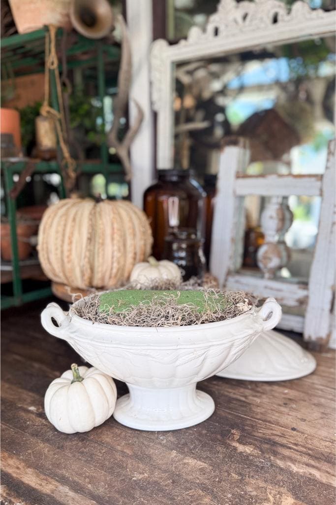 A white ceramic tureen filled with moss and twigs sits on a rustic wooden table next to a small white pumpkin, with a large beige pumpkin and vintage decor in the background.