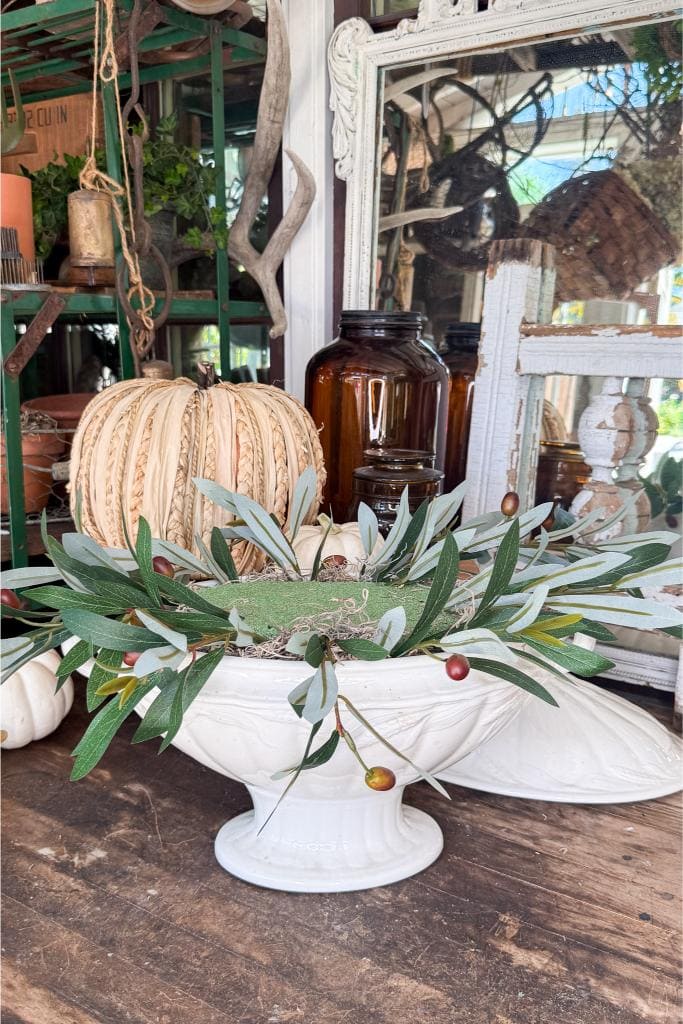 A white pedestal bowl holds faux greenery and a decorative cream pumpkin, set on a rustic wooden table. Amber glass bottles and vintage mirrors are in the background, creating a cozy, autumnal display.