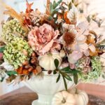A white ceramic vase holds an autumn floral arrangement with hydrangeas, dried leaves, and pink roses. Small white pumpkins are nestled among the flowers and placed beside the vase, all set on a wooden surface.