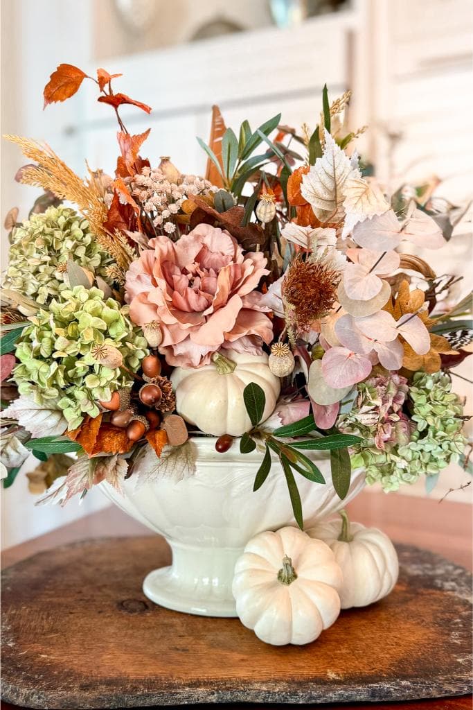 A white ceramic vase holds an autumn floral arrangement with hydrangeas, dried leaves, and pink roses. Small white pumpkins are nestled among the flowers and placed beside the vase, all set on a wooden surface.