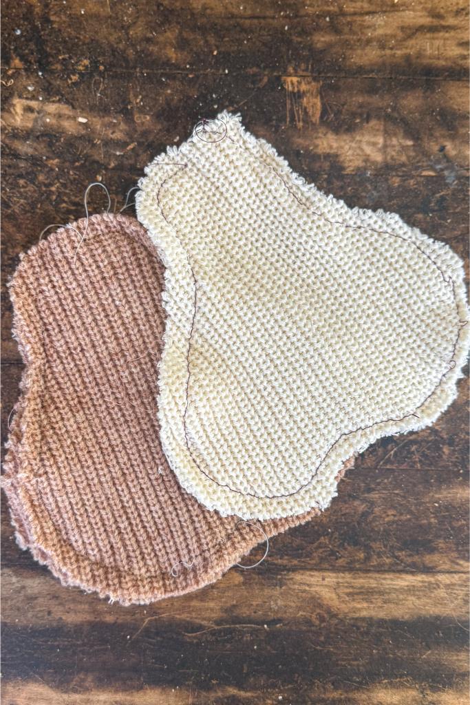 DIY Sweater Pears- irregularly shaped, textured fabric pads—one tan and one off-white—lying on a rustic wooden surface. The stitching and rough edges of the pads are visible.