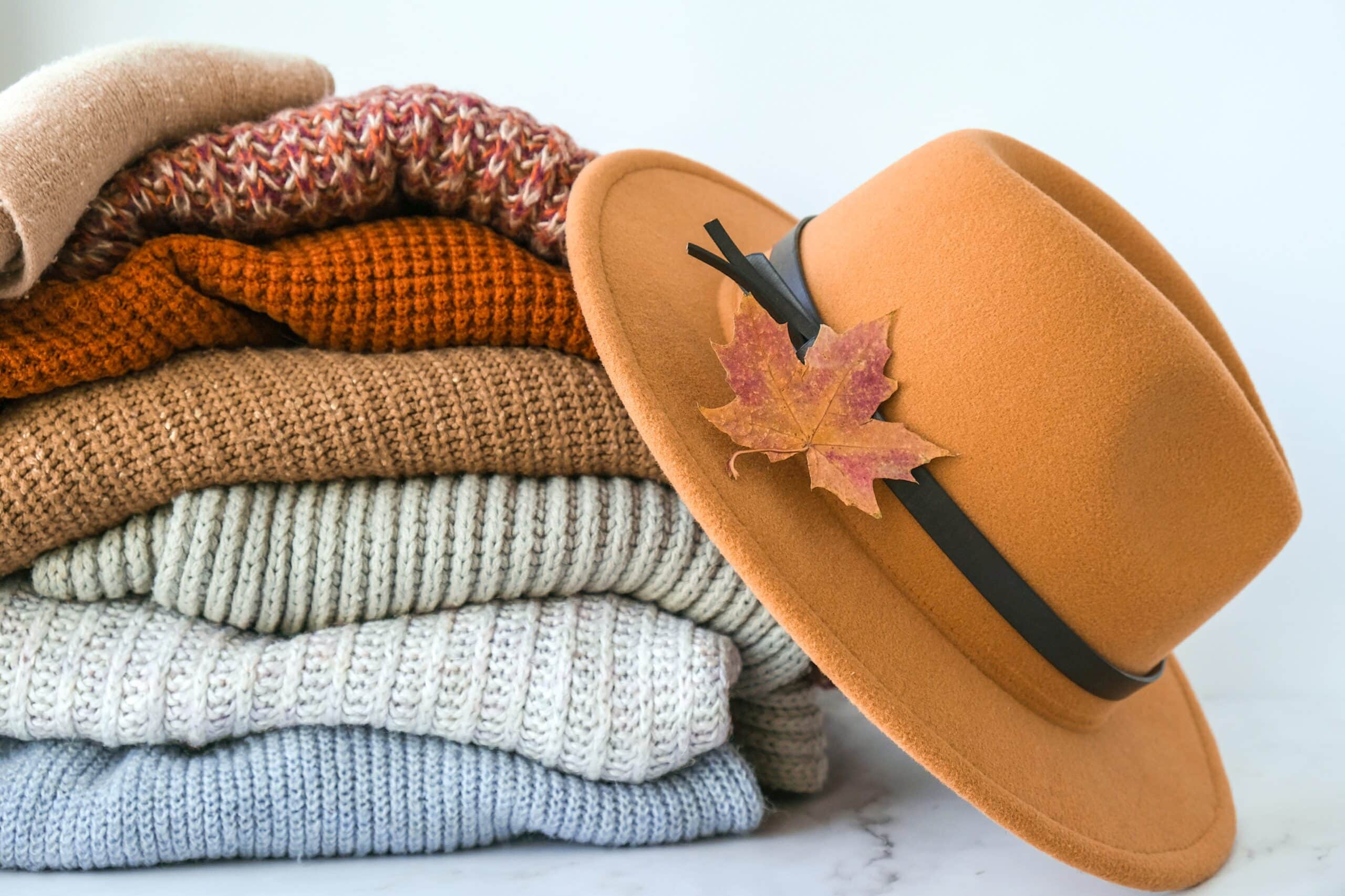 A stack of cozy, folded knit sweaters in various colors sits next to a tan felt hat decorated with a dried autumn leaf—and playful DIY Sweater Pears add a charming seasonal touch.