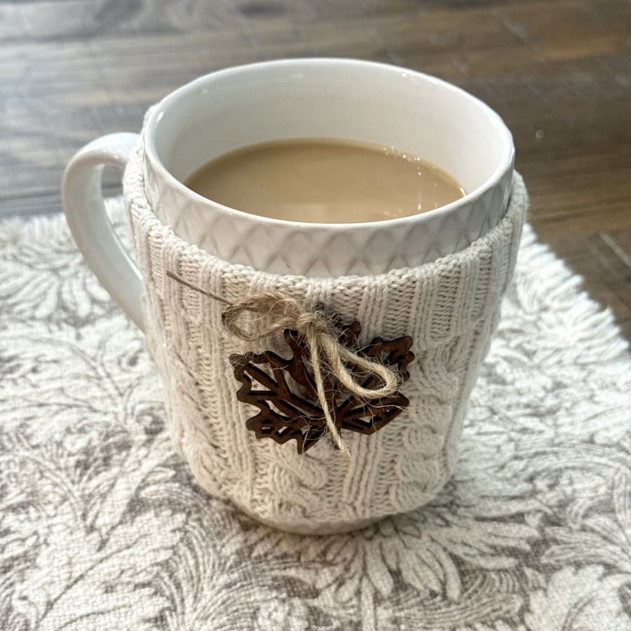 A white mug filled with coffee sits on a patterned surface, wrapped in a cozy knitted cover and decorated with a small wooden leaf and twine bow.