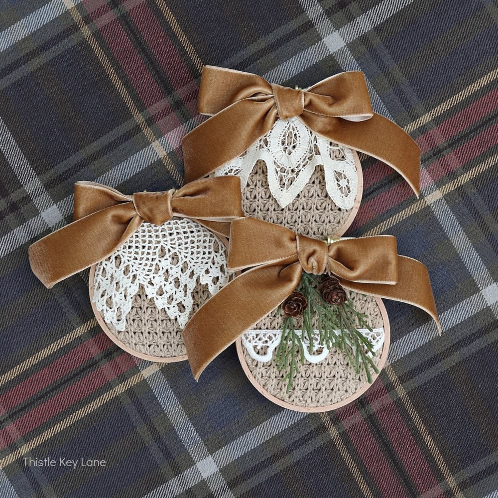 Three round ornaments decorated with burlap, lace, and brown velvet bows rest on a plaid fabric background. One ornament has mini pinecones and greenery, while the others feature intricate lace patterns.