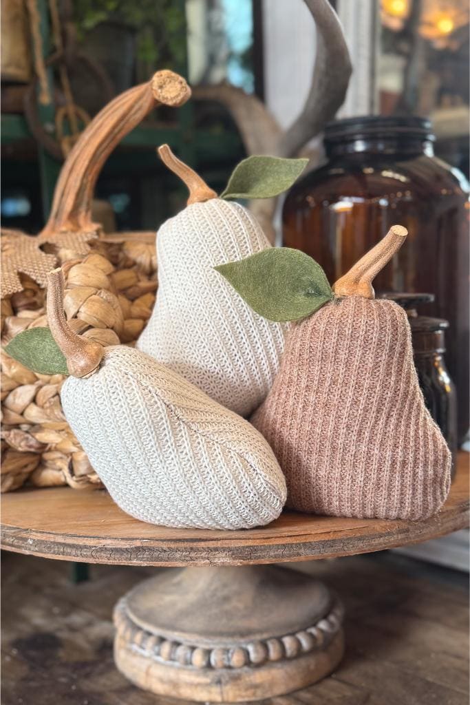 Three knitted fabric pears with felt leaves and twisted stems sit on a wooden tray. The pears are in beige and light brown tones, creating a cozy, rustic decoration. Dark glass jars and baskets are blurred in the background.