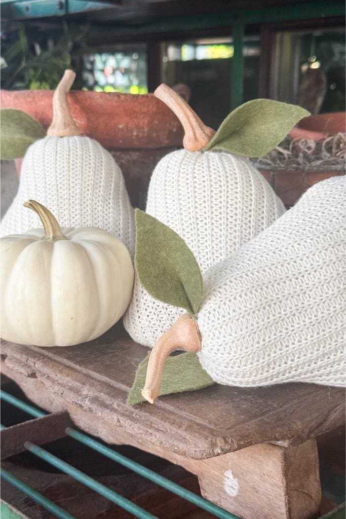 Three pear-shaped objects with knitted white covers and felt leaves sit next to a small white pumpkin on a wooden surface, with a terracotta pot and greenery in the background.