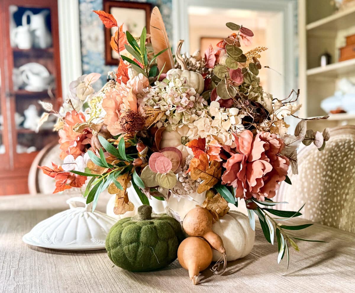 A fall-themed floral arrangement with orange, cream, and green flowers, dried leaves, and pumpkins sits on a wooden table in a DIY fall centerpiece with thrifted container, surrounded by decorative white dishes and a green velvet pumpkin.