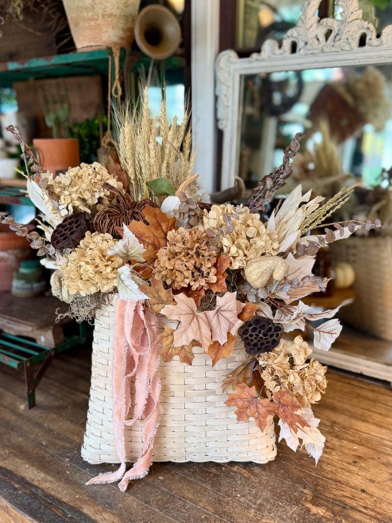A white woven basket filled with dried flowers, wheat, fall leaves, and textured pods sits on a rustic wooden table. Pink ribbon drapes from the arrangement, with vintage decor and a mirror in the background.