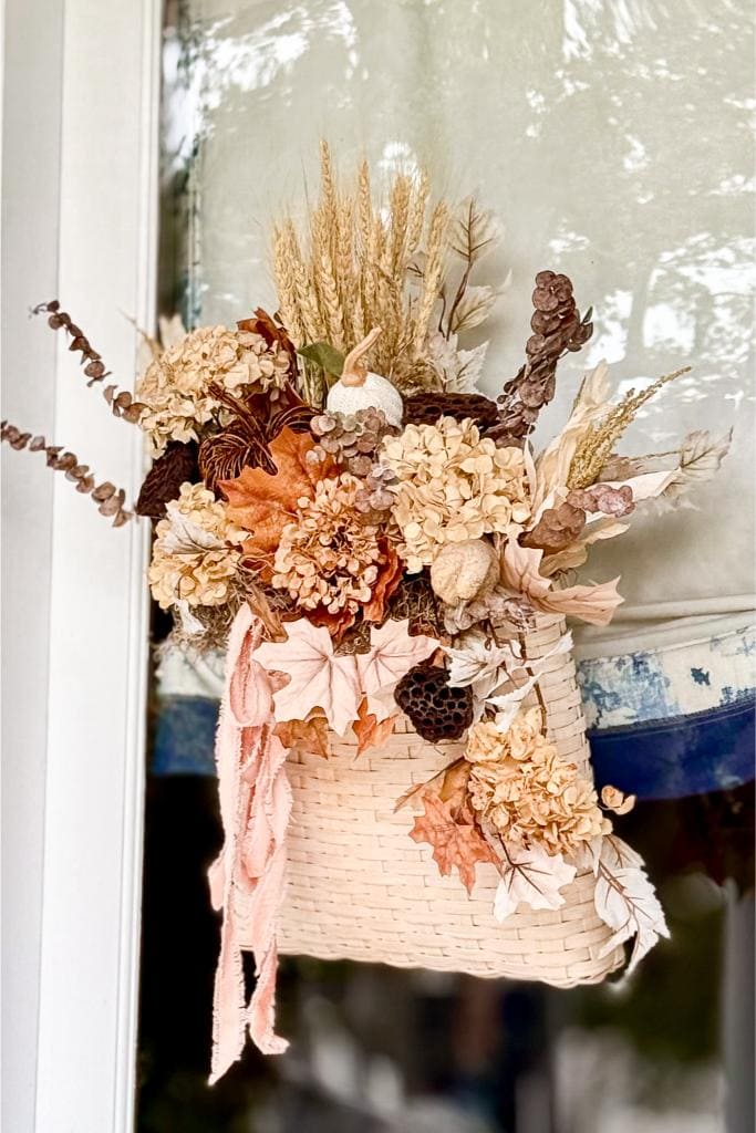 A woven basket filled with dried flowers, leaves, pinecones, and grasses hangs on a door. The arrangement includes neutral tones and a small pumpkin, evoking an autumn or fall theme.